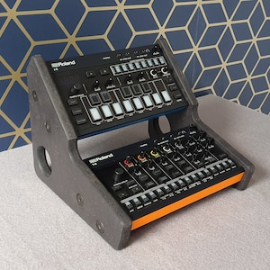 May include: A black and orange double-tiered stand for two Roland electronic music instruments. The stand is made of wood and has a triangular shape. The top tier is for a Roland TR-8S Rhythm Composer and the bottom tier is for a Roland MC-101 Groovebox.