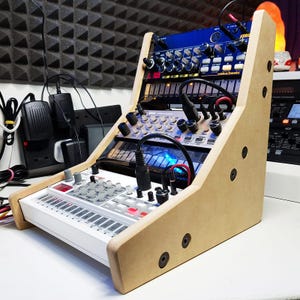 Peut inclure: Un support en bois avec trois synthétiseurs de musique électroniques. Le synthétiseur du haut est bleu et porte l'inscription "Korg Electribe 2 Sampler". Le synthétiseur du milieu est beige et porte l'inscription "MFB Synth II". Le synthétiseur du bas est blanc et a des boutons rouges et noirs.