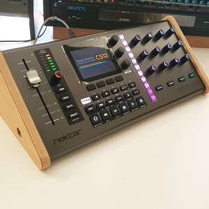 Pode incluir: Um controlador de produção musical cinza com um suporte de madeira. O controlador tem uma tela que exibe "PANORAMA CS12" e uma linha de botões com luzes coloridas.