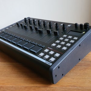 Puede incluir: Un sampler Roland S-2400 negro con un borde plateado. El sampler tiene una variedad de botones y perillas, incluida una pequeña pantalla LCD.