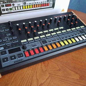 Puede incluir: Una caja de ritmos Rhythm Designer negra y gris con paneles laterales de madera. Cuenta con numerosas perillas, botones y almohadillas de colores. La pantalla muestra una lectura digital. Una caja con un diseño similar está en el fondo.
