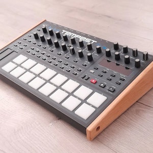 May include: A black Tempest drum machine with a wooden stand. The drum machine has a grid of 16 pads and a row of knobs and buttons.
