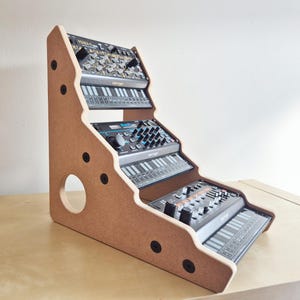May include: A wooden stand with three tiers for holding electronic music synthesizers. The stand is made of light brown wood and has a rounded edge. The stand is angled to the right and has a hole in the bottom left corner. The stand is holding three synthesizers, all of which are black and have a variety of knobs and buttons.