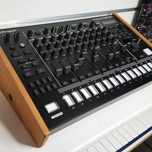 Puede incluir: Caja de ritmos Roland TR-8S negra con paneles laterales de madera. Cuenta con numerosas perillas, controles deslizantes y botones, junto con una fila de teclas blancas. Una caja de sintetizador Waldorf Streichfett es visible en el fondo.