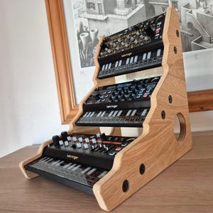 May include: A wooden stand with three tiers for holding electronic musical instruments. The stand is made of light brown wood and has a natural finish. The stand is designed to hold three synthesizers, each with a different colour scheme. The top synthesizer is black with gold accents, the middle synthesizer is black with blue accents, and the bottom synthesizer is black with orange accents. The stand is a great way to organise and display your musical instruments.