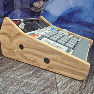 May include: A wooden stand for an Akai MPC-3000 music production centre. The stand is made of light-coloured wood and features a slanted design. The MPC-3000 has a grey and black colour scheme with various buttons and knobs.