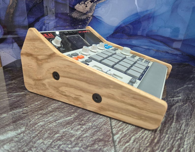 May include: A wooden stand for an Akai MPC-3000 music production centre. The stand is made of light-coloured wood and features a slanted design. The MPC-3000 has a grey and black colour scheme with various buttons and knobs.