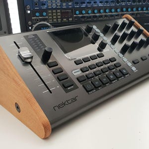 Puede incluir: Un controlador de producci&oacute;n musical negro con un soporte de madera. El controlador tiene una pantalla grande, botones, botones y un fader. El texto "nektar" est&aacute; impreso en la parte frontal del controlador.