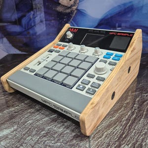 May include: An Akai MPC Sample music production centre with a light wood frame. The device has a grey and white interface with numerous buttons, pads, and knobs. The text "MPC SAMPLE" is visible on the front.