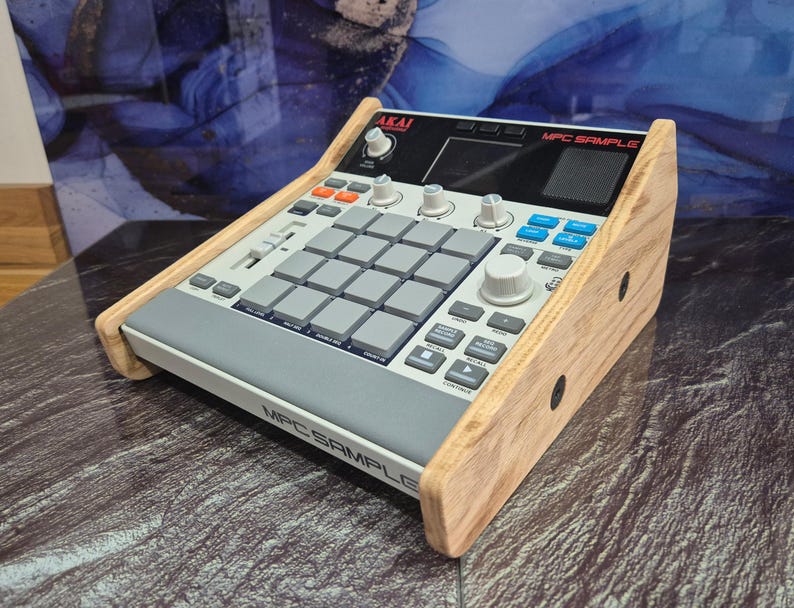May include: An Akai MPC Sample music production centre with a light wood frame. The device has a grey and white interface with numerous buttons, pads, and knobs. The text "MPC SAMPLE" is visible on the front.