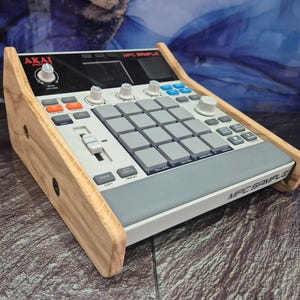 May include: An Akai MPC Sample music production device with a wooden frame. The device has grey pads, orange and blue buttons, and white knobs. The text "MPC SAMPLE" is visible on the front.