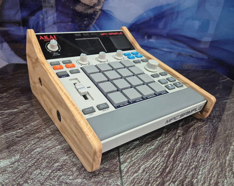 May include: An Akai MPC Sample music production device with a wooden frame. The device has grey pads, orange and blue buttons, and white knobs. The text "MPC SAMPLE" is visible on the front.