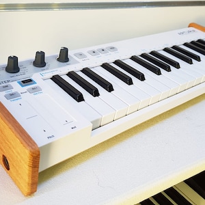 Puede incluir: Teclado MIDI blanco de 32 teclas con paneles laterales de madera. El teclado tiene varios botones y perillas para controlar la música.