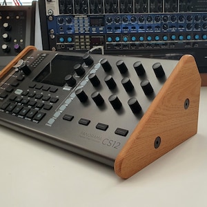 May include: A silver synthesizer with a wooden stand. The synthesizer has a black display screen, a grid of buttons, and a row of knobs. The text "Panorama CS12" is printed on the front of the synthesizer.