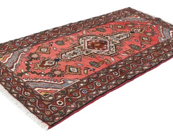 May include: A rectangular area rug featuring a vibrant coral-red field with a central geometric medallion in cream, navy, and brown. The border displays a repeating wave pattern in dark red, accented with small floral and geometric motifs.