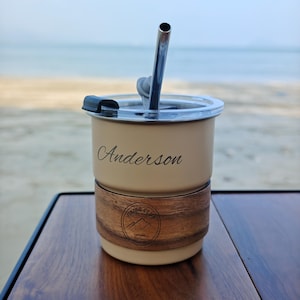 May include: A beige insulated tumbler with a stainless steel straw and lid, resting on a wooden base. The name "Anderson" is elegantly scripted on the tumbler. A wooden band with a "Hiking Club" logo adds a rustic touch.
