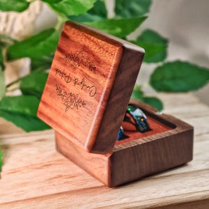 May include: A wooden ring box with a hinged lid, engraved with floral designs and the names "Cindy & Patrick." The box is open, revealing a ring with blue and green accents. The box is made of dark wood.