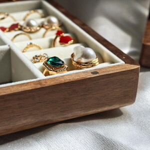 May include: A wooden jewelry box with a light-colored interior, showcasing various rings. The rings feature gemstones, pearls, and gold settings. One ring has a large emerald-colored stone, and another has a large pearl.