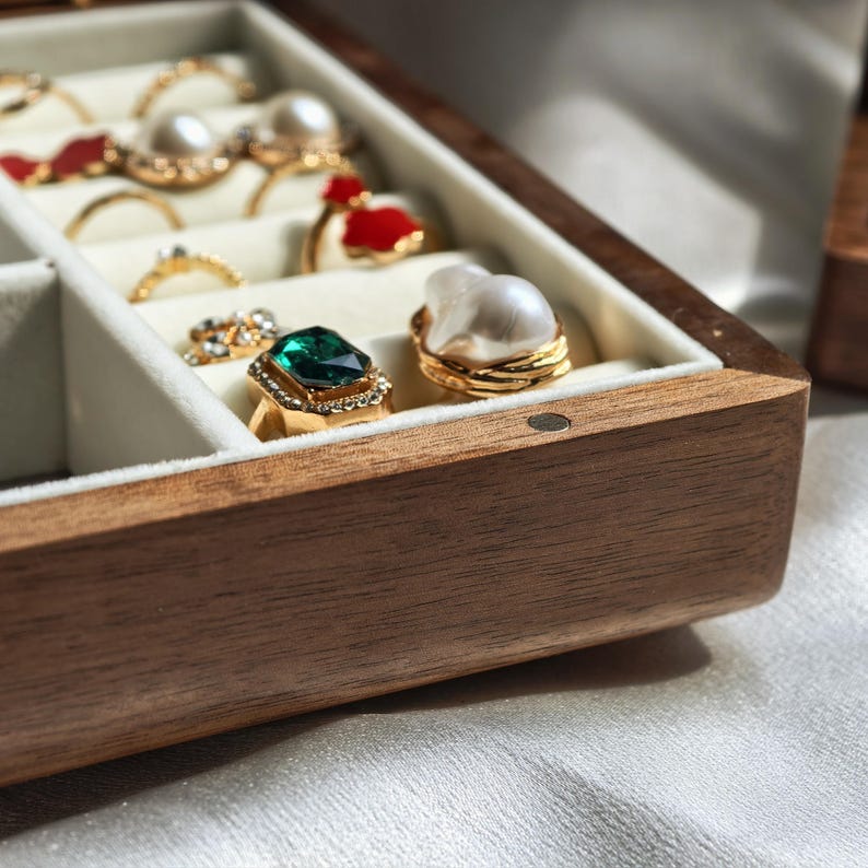 May include: A wooden jewelry box with a light-colored interior, showcasing various rings. The rings feature gemstones, pearls, and gold settings. One ring has a large emerald-colored stone, and another has a large pearl.