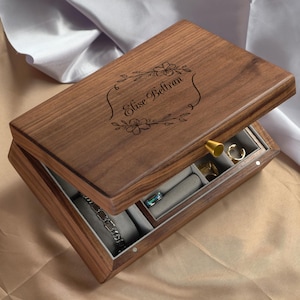 以下が含まれることがあります： 木製のジュエリーボックス。蓋が開いており、灰色の布で裏打ちされたコンパートメントが見えます。ボックスには、花柄のデザインの中に「Elise Beltran」という名前が刻まれています。 金色の留め金が蓋を固定しています。