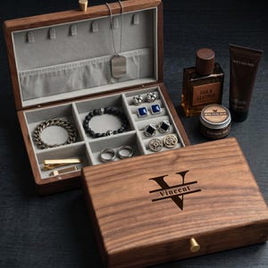 May include: A dark wood jewellery box with a grey velvet interior, featuring the engraved name Vincent and a large letter V on the lid. The box contains a silver chain, a beaded bracelet, a metal chain bracelet, cufflinks, and rings. Nearby are a bottle of Oak & Leather cologne and a jar of facial balm.