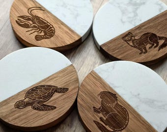 May include: Four round coasters with a two-tone design. Half of each coaster is wood-toned with an engraved animal: a lobster, a weasel, a turtle, and a beaver. The other half is white with a marble pattern.