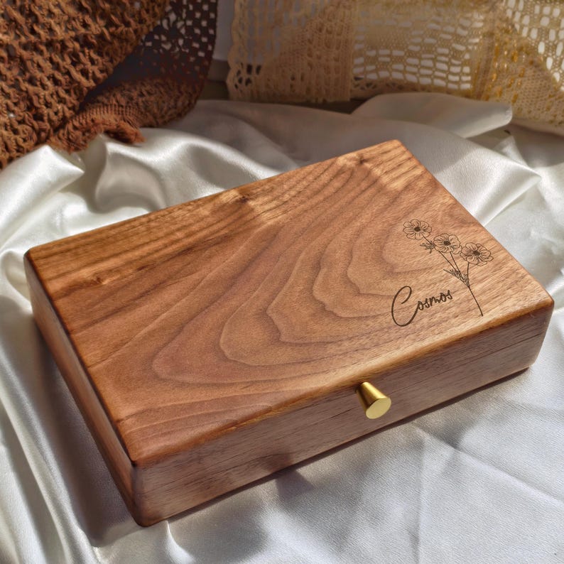 May include: A rectangular wooden box with a natural wood grain finish. The box has a gold-colored knob and a floral design with the word "Cosmos" engraved on the top. The box is sitting on a white satin surface.