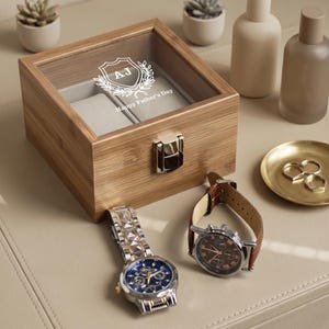 May include: A bamboo watch box with a glass lid, engraved with "AJ" and "Happy Father's Day". Two watches with leather and metal straps are displayed. A small gold dish with rings is also visible.