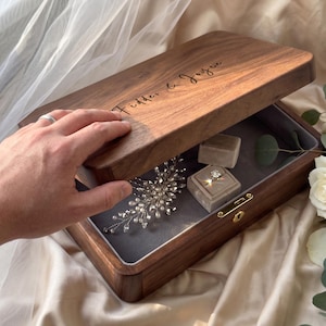May include: A rectangular dark wood keepsake box with a hinged lid engraved with the text Fidder & Joyce. The interior is lined with grey fabric and contains a silver-toned hair accessory with pearls and a ring inside a small velvet box.