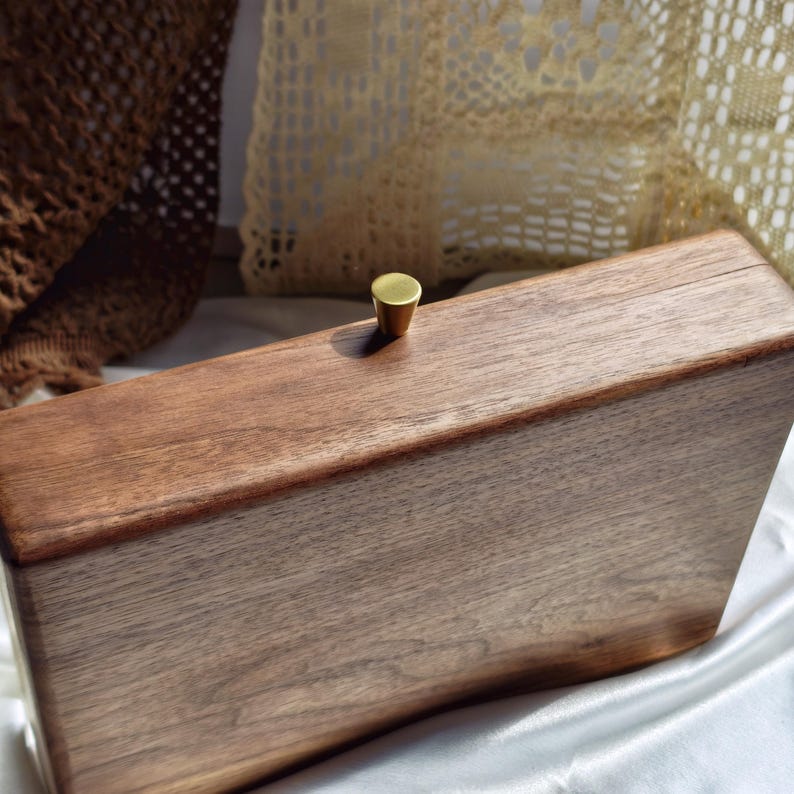 May include: A rectangular wooden clutch with a brass knob. The clutch is made of dark brown wood and has a smooth, polished finish. The brass knob is small and round. The clutch is sitting on a white satin surface.