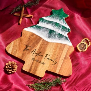 May include: A wooden, Christmas tree-shaped serving board with a green and white resin design. The board is engraved with "The Austin Family" and the date "08.08.2024". Cinnamon sticks, rosemary, a pinecone, and dried lemon slices are arranged around the board.