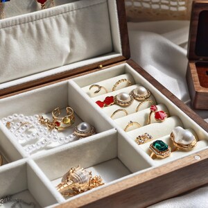 May include: A wooden jewelry box with a light-colored interior, filled with various rings, necklaces, and earrings. The jewelry includes pearls, gold, and gemstones. The box is open, revealing multiple compartments.