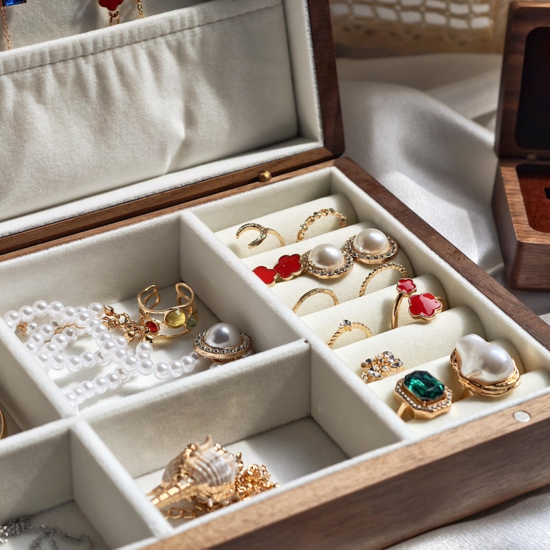 May include: A wooden jewelry box with a light-colored interior, filled with various rings, necklaces, and earrings. The jewelry includes pearls, gold, and gemstones. The box is open, revealing multiple compartments.