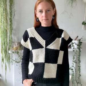 Puede incluir: Un chaleco de punto con estampado de tablero de ajedrez en negro y crema con cuello en V. El chaleco se lleva sobre una camisa negra de manga larga. El estilo general es moderno e informal.