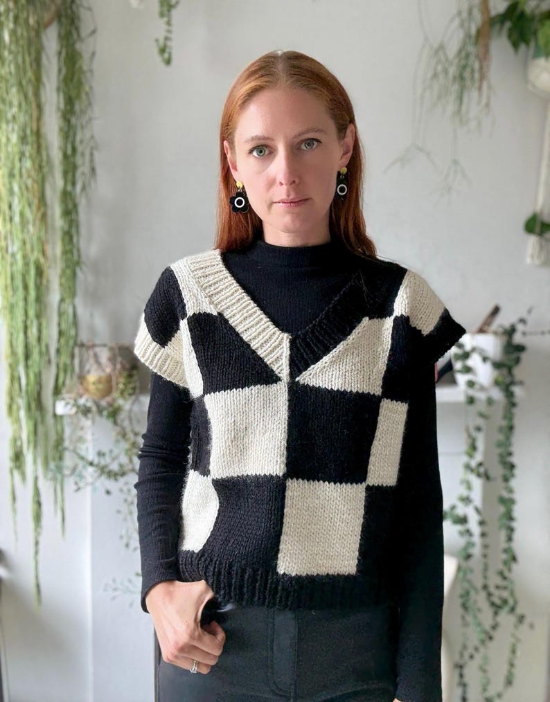 May include: A black and cream checkerboard pattern sweater vest with a V-neck. The vest is worn over a black long-sleeved top. The style is modern and casual.