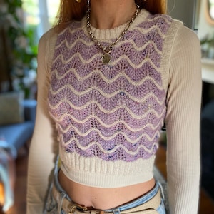 Puede incluir: Un chaleco corto sin mangas con un patrón ondulado de color lavanda y crema. El chaleco está superpuesto a una camisa crema de manga larga y acanalada. Un collar plateado y pendientes florales completan el look. La persona lleva un cinturón de color claro y vaqueros azules.