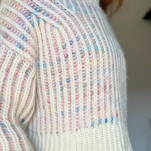 May include: A cream-coloured, ribbed knit jumper with a high neck. The jumper has vertical stripes with pastel pink, blue, and yellow flecks. The sleeves are long, and the overall style is cosy and textured.