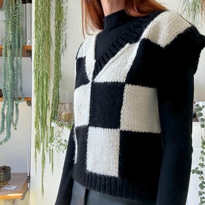 May include: A black and cream-coloured checkerboard knit vest with a V-neck. The sleeveless sweater is made of a chunky knit material. The vest is worn over a black turtleneck.