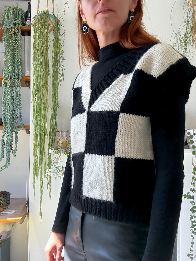 May include: A black and cream-coloured checkerboard knit vest with a V-neck. The sleeveless sweater is made of a chunky knit material. The vest is worn over a black turtleneck.