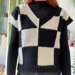 May include: A knitted vest featuring a black and cream checkerboard pattern and a V-neck. The sleeveless top is worn over a black long-sleeved garment. The design of the vest showcases a geometric pattern of alternating black and cream squares.