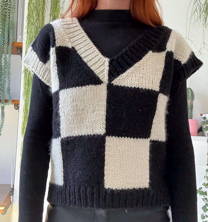 May include: A knitted vest featuring a black and cream checkerboard pattern and a V-neck. The sleeveless top is worn over a black long-sleeved garment. The design of the vest showcases a geometric pattern of alternating black and cream squares.