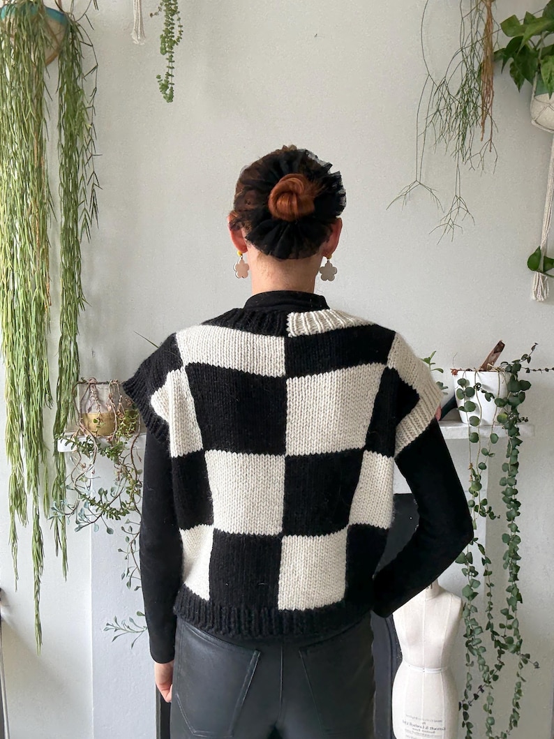 May include: A black and white checkerboard knit vest. The sleeveless sweater has a high neckline and a classic checkerboard pattern. The vest is worn over a black long-sleeved shirt and paired with black leather trousers. The model is facing away from the camera.