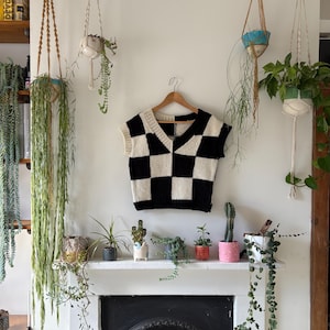 May include: A black and cream-coloured checkerboard pattern knit vest hangs on a wooden hanger. The vest has a V-neck and is sleeveless. The background includes a white wall, a fireplace mantel with potted plants, and hanging plants.