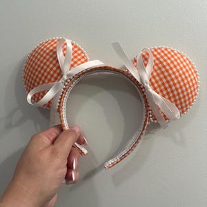 May include: Orange and white gingham pattern mouse ear headband with white satin bows. The headband is trimmed with white pearls and a white crocheted edge. A fun accessory for themed events.