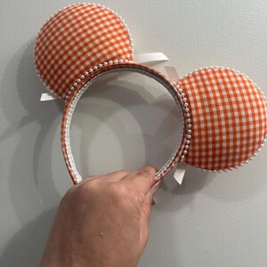 May include: Orange and white gingham pattern mouse ear headband. The ears are round and trimmed with white pearls. The headband is white with orange gingham trim and white ribbons.