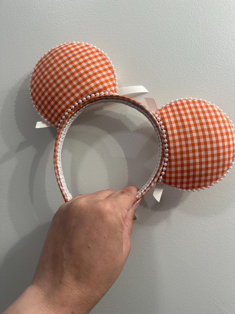 May include: Orange and white gingham pattern mouse ear headband. The ears are round and trimmed with white pearls. The headband is white with orange gingham trim and white ribbons.