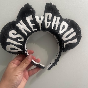 May include: Black sequined headband with mouse ears. The ears have the text "DISNEY GHOUL" in white block letters. The headband is held by a hand against a light gray background.