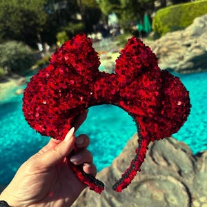 May include: Red sequined Minnie Mouse ears headband. The headband is made of a black fabric with a red sequined bow on top.