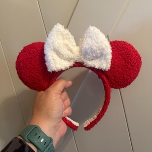May include: Red fuzzy Minnie Mouse ears with a white fuzzy bow. The headband is trimmed with white and cream.