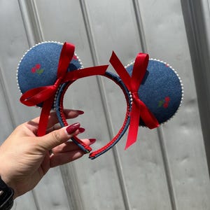 May include: Denim-style mouse ears headband with red bows and cherry embroidery. The ears are outlined with white pearls, and the headband is red and blue. A fun accessory for themed events.